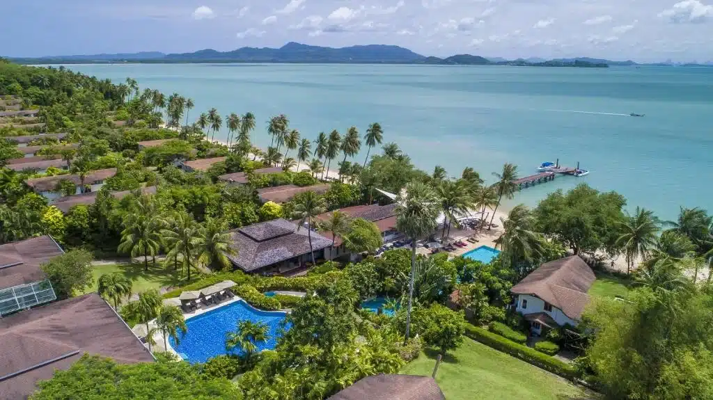 Aerial view of one of the best luxury beachfront resorts in Phuket by a calm, blue sea. The resort features several bungalows with brown roofs, two rectangular swimming pools, and lush green trees. A sandy beach stretches along the shoreline, with a wooden pier extending into the water. Hills are visible on the horizon.