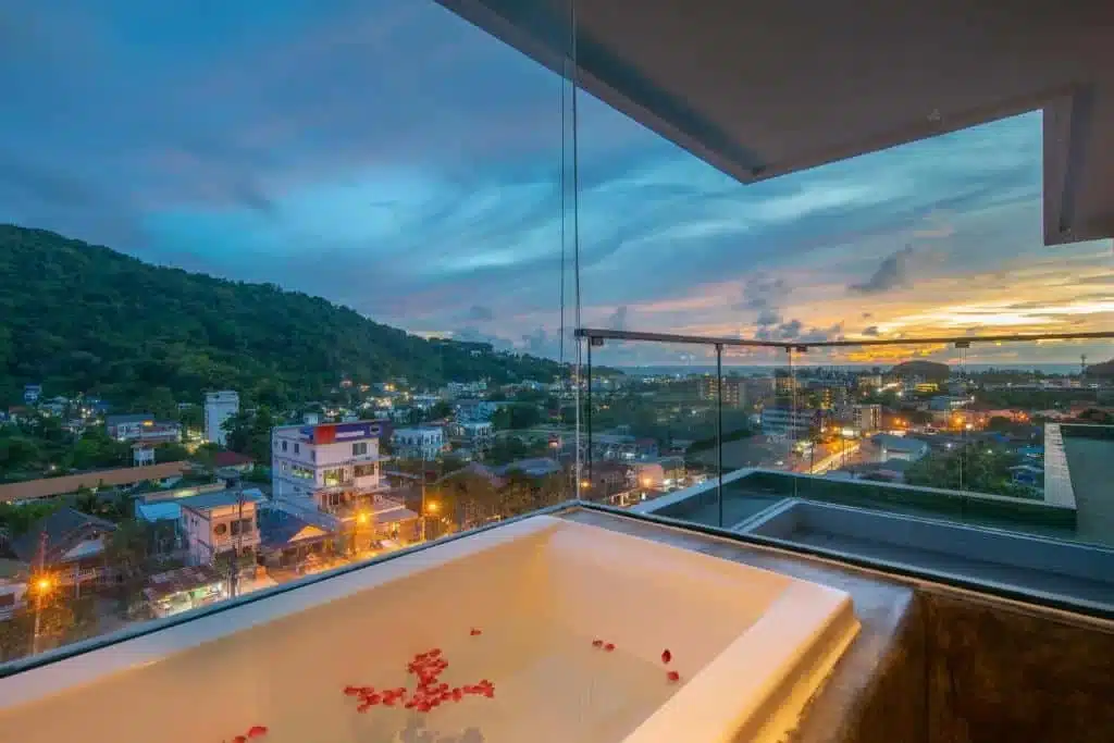 An outdoor bathtub with scattered flower petals sits on a balcony overlooking a cityscape at dusk, including the majestic Big Buddha Phuket. The view features illuminated buildings, lush green hills, and a partly cloudy sky with the sun setting in the background. The scene is serene and picturesque.