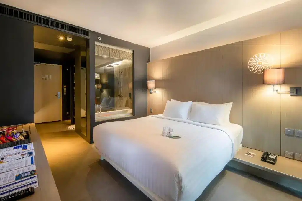 A modern hotel room featuring a neatly made double bed with white linens, positioned against a light wood panel wall with two mounted lamps. The room includes a glass-walled bathroom on the left, a wooden desk with snacks and brochures in the foreground highlighting nearby attractions like Big Buddha Phuket, and minimalistic decor throughout.