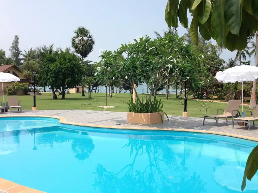 A serene outdoor scene features a clear blue swimming pool surrounded by lounge chairs with white umbrellas. Adjacent to the pool is a lush green grassy area with trees and shrubs, leading to a view of the ocean in the background. The sky is clear, indicating a sunny day at one of the best hotels in Phuket near the beach.