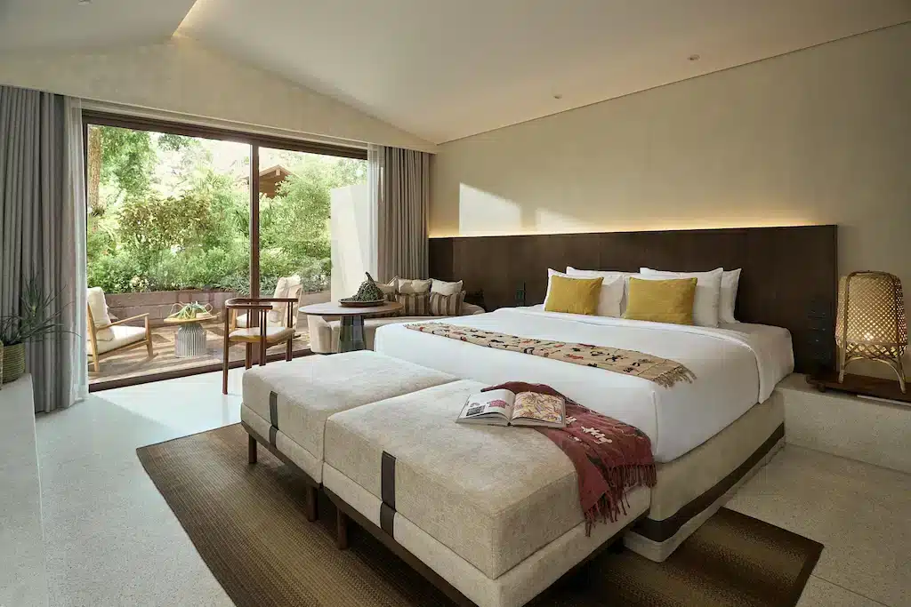 Luxurious hotel room at one of the best hotels in Phuket near the beach, featuring a large bed with white linens and mustard accent pillows, flanked by two beige ottomans. The room includes a wooden chair, small table, cushioned sofa, and opens to a patio with outdoor seating and greenery visible through glass sliding doors.