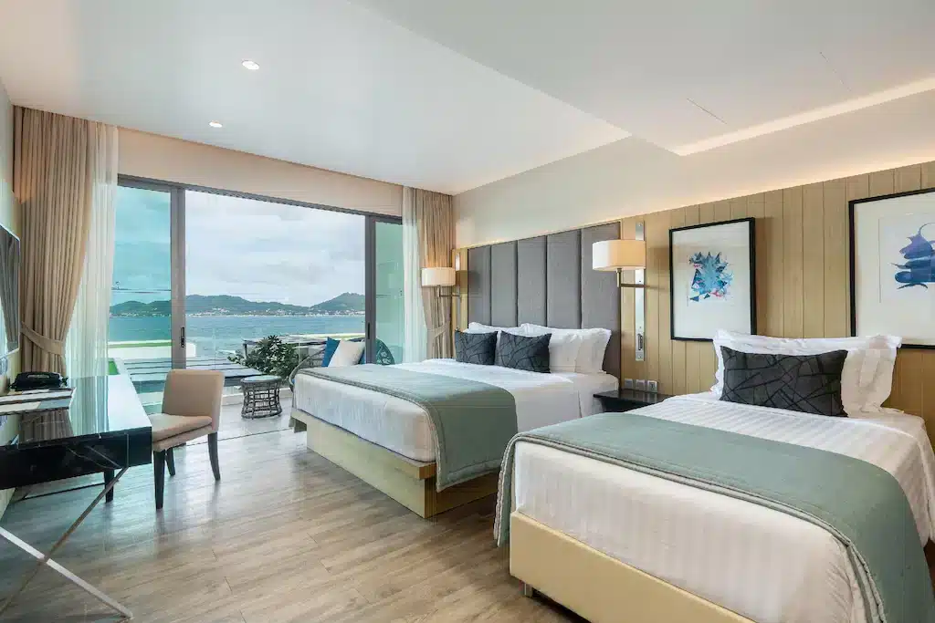 A modern, well-lit hotel room with two large beds, each dressed in white linens and green throws. A desk and chair sit to the left, while large glass doors offer an ocean view with mountains in the distance. The room features light wood flooring, neutral-toned walls, and decorative wall art—exemplifying one of the best hotels in Phuket near the beach.