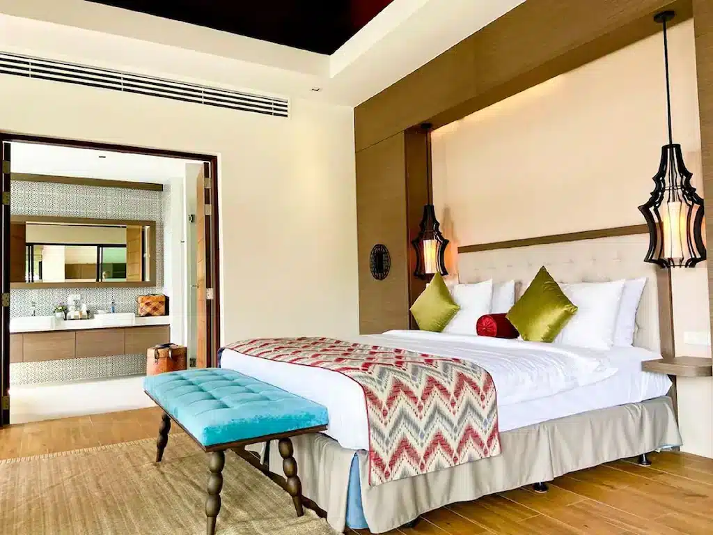 A cozy hotel room features a large, neatly made bed with green and red throw pillows and a zigzag-patterned blanket. A blue cushioned bench sits at the foot of the bed. Unique hanging lamps flank the bed, and the room opens up to a modern, well-lit bathroom with a large mirror, mirroring the serenity of Big Buddha Phuket.