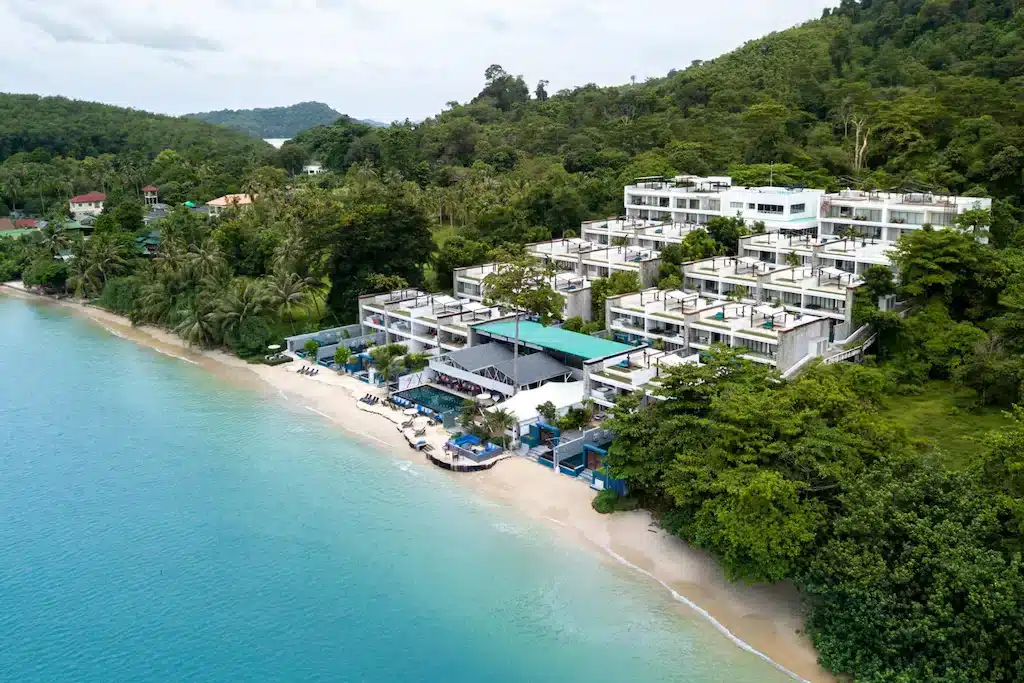 An oceanfront resort with multiple white buildings nestled on a lush, green hillside. The resort features modern architecture with terraced balconies. Below, a pristine sandy beach meets turquoise waters. Palm trees and dense vegetation frame the beach, making it one of the best hotels in Phuket near the beach.