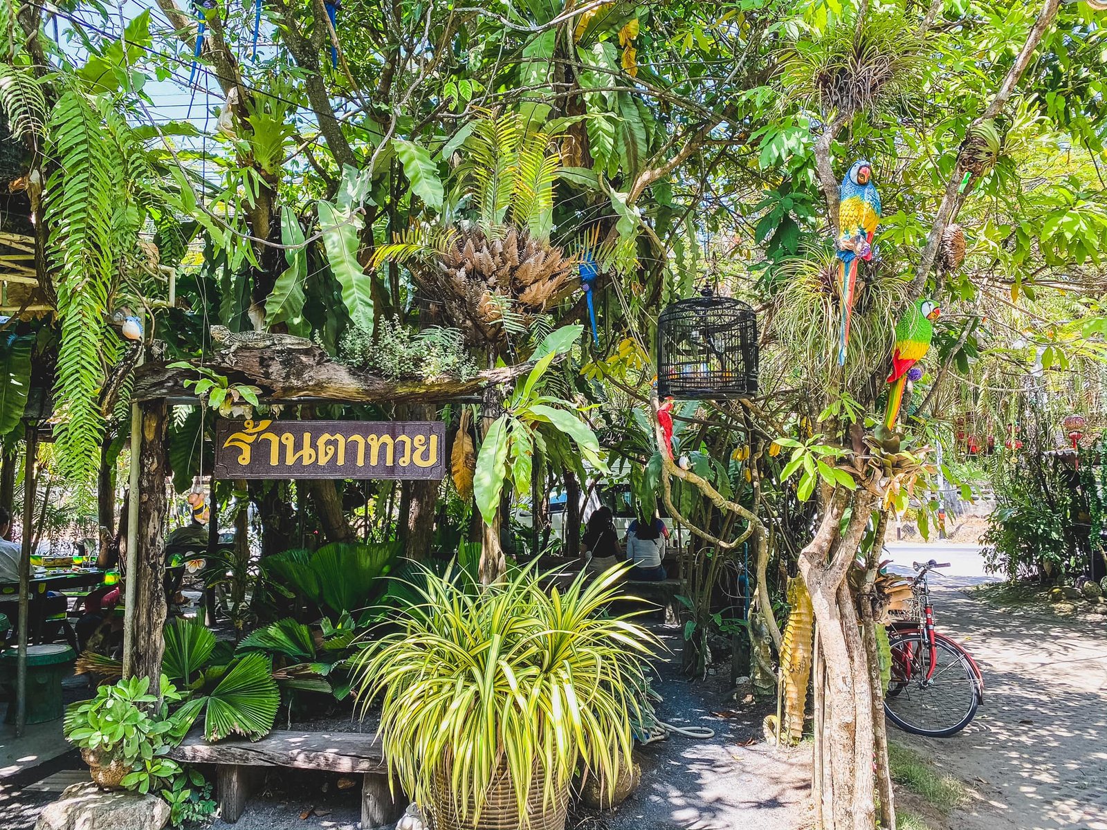 A lush garden restaurant features a wooden sign with Thai writing. The space is adorned with verdant plants, hanging ferns, and colorful tropical decorations reminiscent of Sarasin Love Bridge. A red bicycle leans against a tree, and two vibrant parrot figurines add a splash of color to the green surroundings.