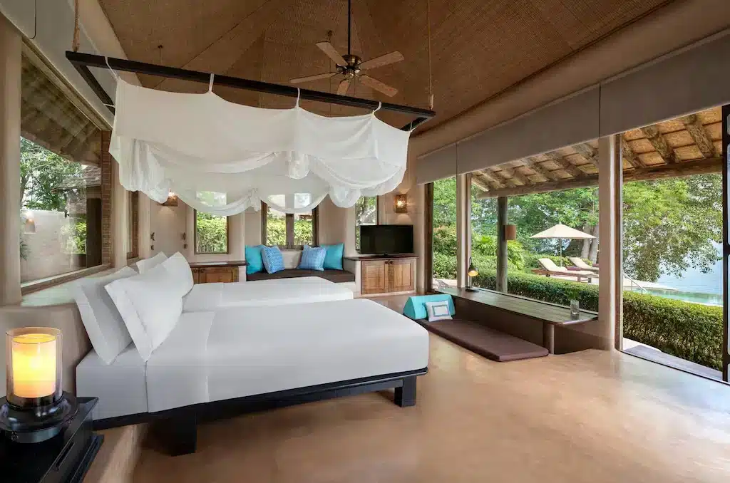 Luxurious bedroom with a scenic view of a garden and lake through large windows. The room features two white canopy beds, a wooden ceiling fan, a cushioned window seat, and a TV. Outside, there's a deck with lounge chairs and an umbrella reminiscent of luxury beachfront resorts in Phuket. A lit candle adds to the cozy ambiance.