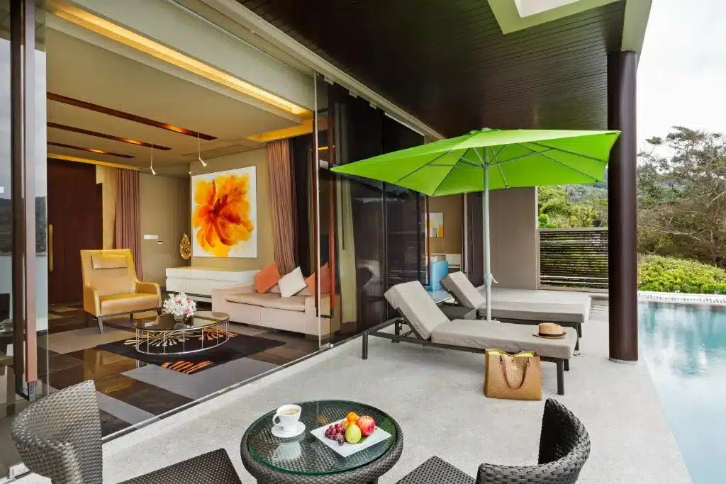 A luxurious hotel room with an open sliding glass door leading to a private patio. The patio features two lounge chairs under a large green umbrella, a wicker table set with fruit, and a coffee cup. A swimming pool lies adjacent to the patio, and a tropical landscape is visible in the background at one of the luxury beachfront resorts in Phuket.