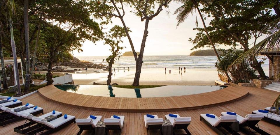 A luxurious beachfront pool area with wooden decking and white lounge chairs topped with blue towels overlooks a serene beach. Tall trees frame the foreground. People can be seen enjoying the gentle waves and golden sand under a clear sky in the background, typical of luxury beachfront resorts in Phuket.