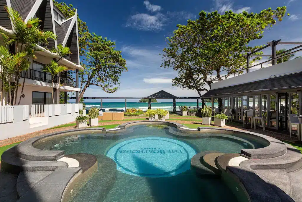 A scenic view of a resort featuring a uniquely shaped pool with "The Boathouse Phuket" inscribed at its center. Surrounding the pool, there are modern white buildings with balconies, outdoor dining areas, and lush greenery. Beyond the resort, one of the best hotels in Phuket near the beach offers a picturesque beach and sea under a clear blue sky.