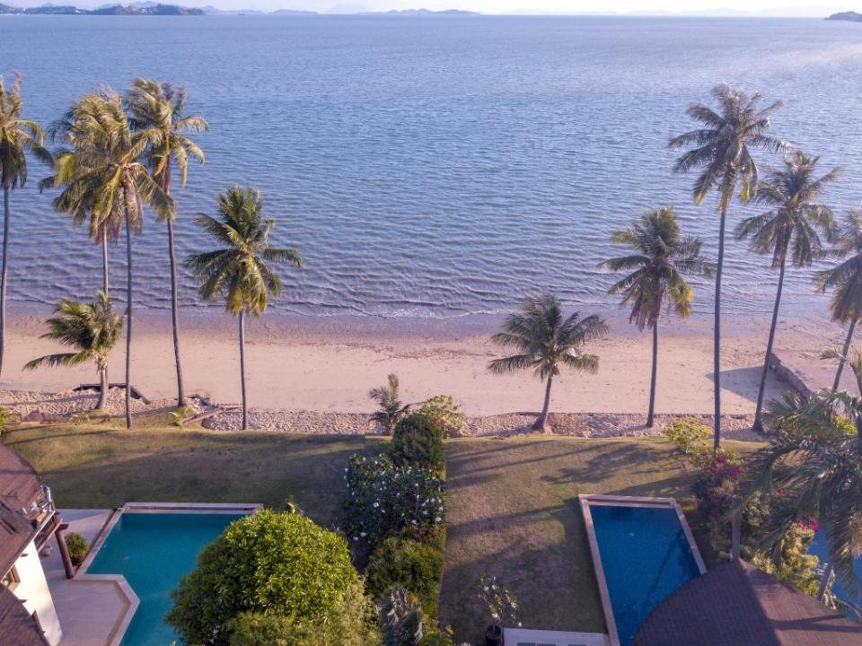 A serene beachfront view at one of the best hotels in Phuket near the beach features tall palm trees lining a sandy shoreline, with gentle ocean waves in the background. Two swimming pools are visible on green lawns adjacent to the beach, surrounded by lush greenery and a large house. The sky is clear and the distant horizon can be seen.