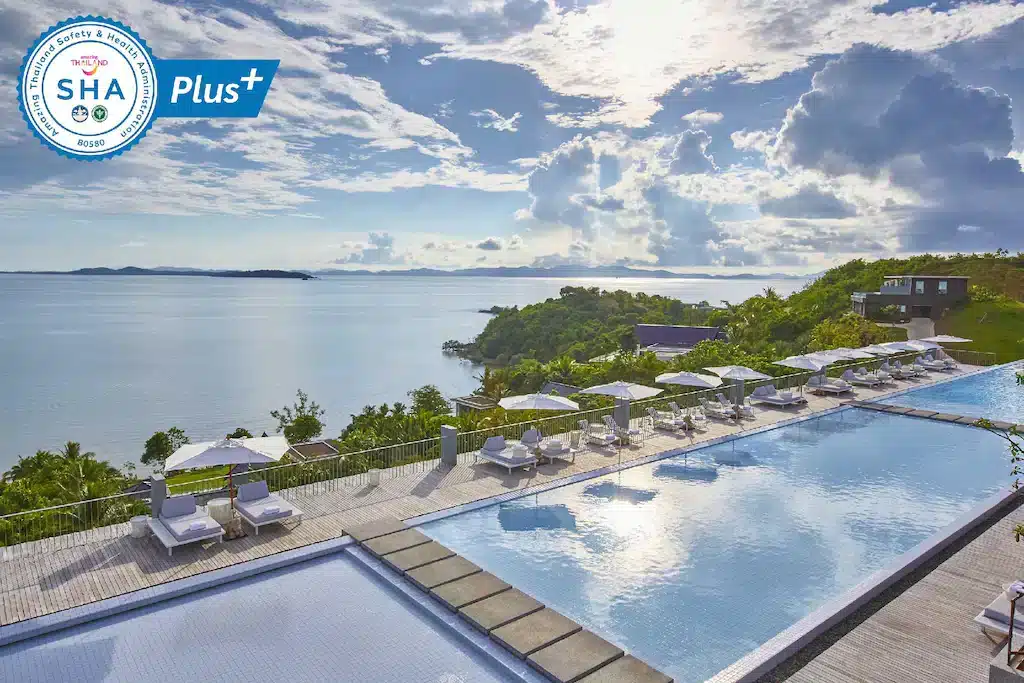 A scenic view of a luxury resort with a large infinity pool overlooking a serene ocean. Lounge chairs with white umbrellas line the poolside on a spacious deck. The sky is partly cloudy, and lush greenery surrounds the area. Among Phuket beachfront resorts, it boasts a "SHA Plus+" certification logo in the top left corner.