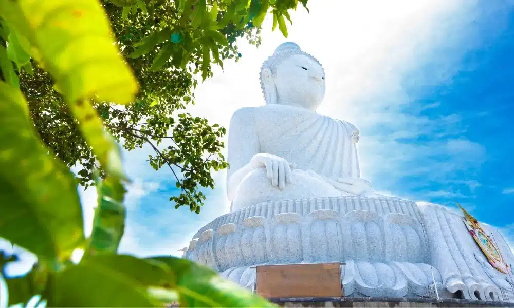 A large, white stone Buddha statue in a seated position, framed by green foliage on the left side. Set against a bright blue sky with wispy clouds, this Big Buddha Phuket monument is captured from a low angle, emphasizing its grandeur and peaceful expression.