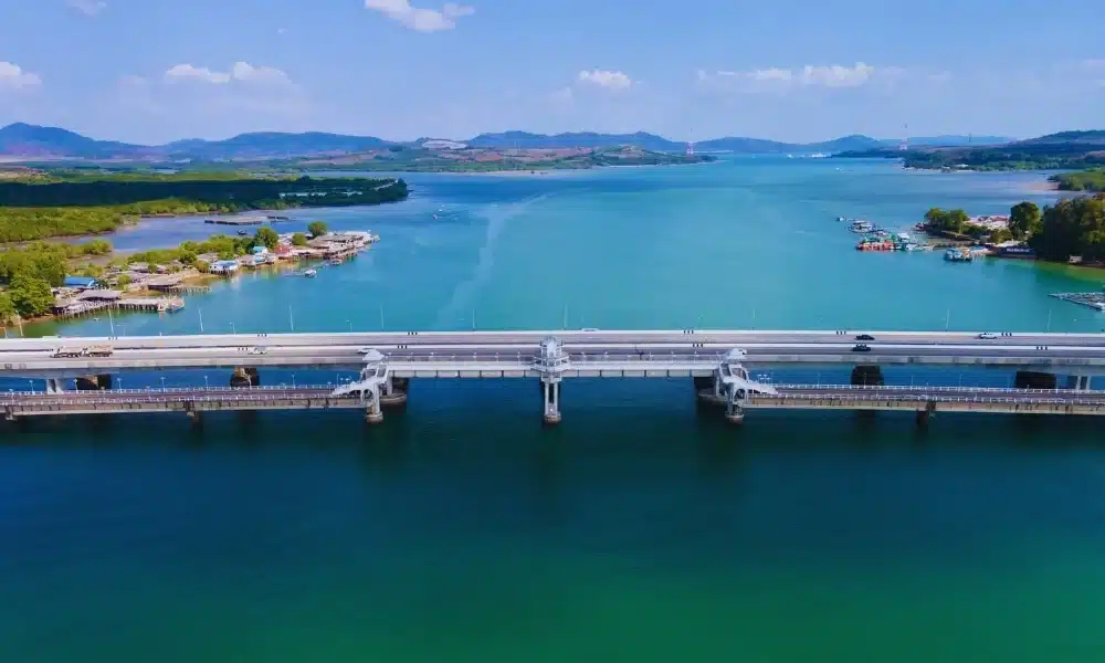 A scenic view of the Sarasin Love Bridge stretching across a calm, turquoise river. The bridge has several supporting pillars and carries light vehicle traffic. On either side of the river, lush green trees and small buildings are visible. In the background, rolling hills extend towards the horizon under a clear blue sky.