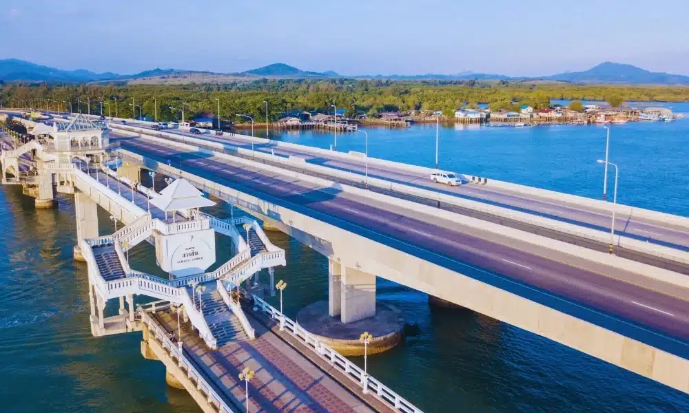 A wide-angle view of the Sarasin Bridge stretching over a large body of water, leading to a forested area and a distant mountain range. A unique two-story observation platform with stairs and a small shelter is visible in the foreground, and several vehicles travel along the bridge under a clear blue sky.