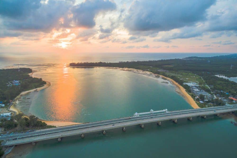 An aerial view of a serene coastal landscape at sunset features the Sarasin Bridge stretching across a wide body of water reflecting the orange and pink hues of the descending sun. The land curves around the water, with green vegetation and scattered buildings lining the shore, while clouds partially cover the sky.