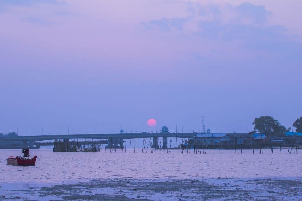 A serene scene of a pinkish sunset over a calm river with a boat and a fisherman in the foreground. The Sarasin Bridge extends across the river, leading to buildings and a tower on the other side. The sky is watercolor-like, blending soft blue and pink hues.