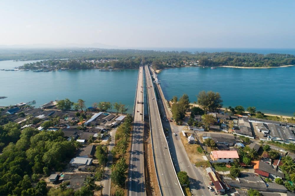 Aerial view of the Sarasin Love Bridge spanning across a body of water, connecting two land masses with residential areas on either side. The bridge has a clear, sunny sky overhead, and the surrounding landscape features clusters of houses, trees, and small roads along the water's edge.