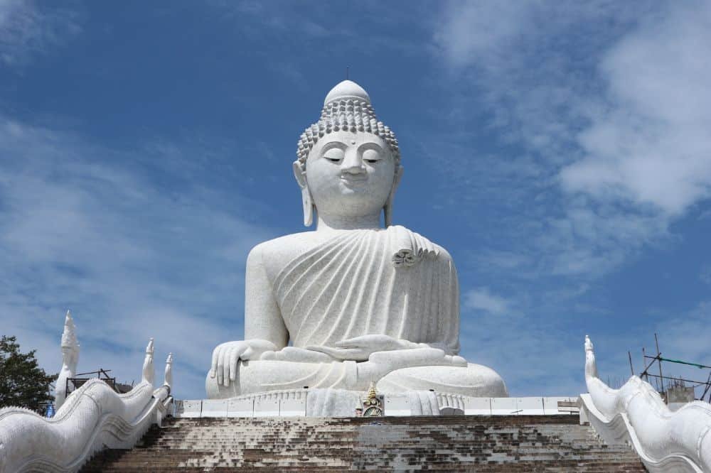 The image shows the Big Buddha Phuket, a large white statue of Buddha in a seated meditation position, with a serene expression. The statue is positioned on a raised platform with staircases adorned with sculpted serpent railings on either side, against a backdrop of a bright blue sky with scattered clouds.