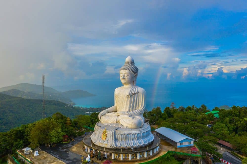 Aerial view of the Big Buddha Phuket statue in Thailand, sitting in a meditative pose on a hilltop. The massive white statue overlooks lush green forests and the blue sea. A rainbow arcs through the sky, adding color to the scene. A few buildings and a telecommunications tower are nearby.