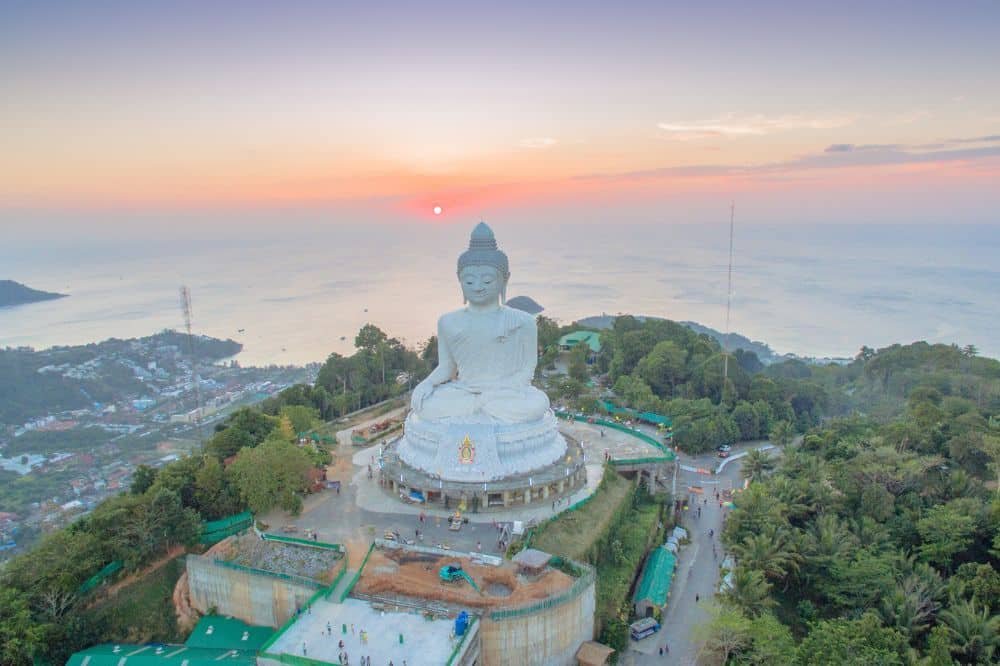 Aerial view of the Big Buddha Phuket statue in Thailand at sunset. The massive white statue sits on a hilltop, surrounded by lush greenery and overlooking the distant ocean. The sky is gradient, transitioning from purple to orange, casting a serene glow over the landscape.