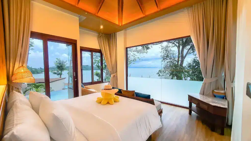 A bright, modern bedroom with large windows offering a panoramic view of the ocean and a lush green landscape, similar to those found in luxury beachfront resorts in Phuket. The room features a neatly made bed with white linens and a yellow towel folded into a decorative shape. The interior is decorated with wooden furniture and warm lighting.