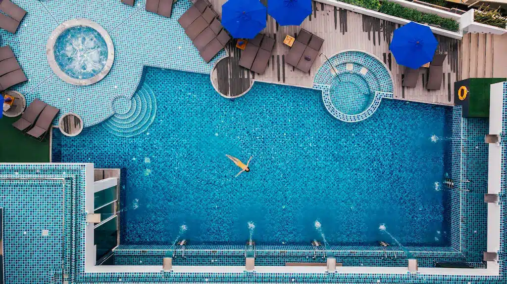 Aerial view of a sparkling blue swimming pool with one person swimming, surrounded by wooden decking with brown lounge chairs and blue umbrellas. Adjacent to the circular hot tub and smaller pool with stairs, you can even spot the iconic Big Buddha Phuket in the distance. Greenery adorns the top right corner.