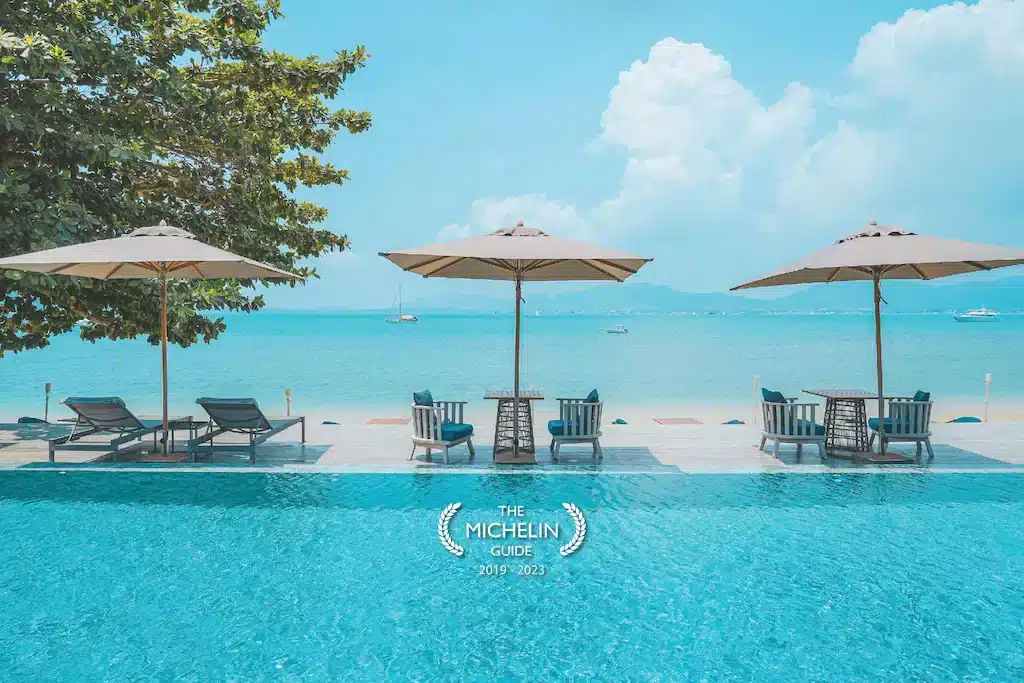 A picturesque Phuket beachfront resort featuring a clear blue swimming pool, sun loungers, and shaded tables with umbrellas overlooking a tranquil sea. Boats float in the distance under a bright sky with fluffy clouds. The image also displays a Michelin Guide logo with the years 2019-2023.