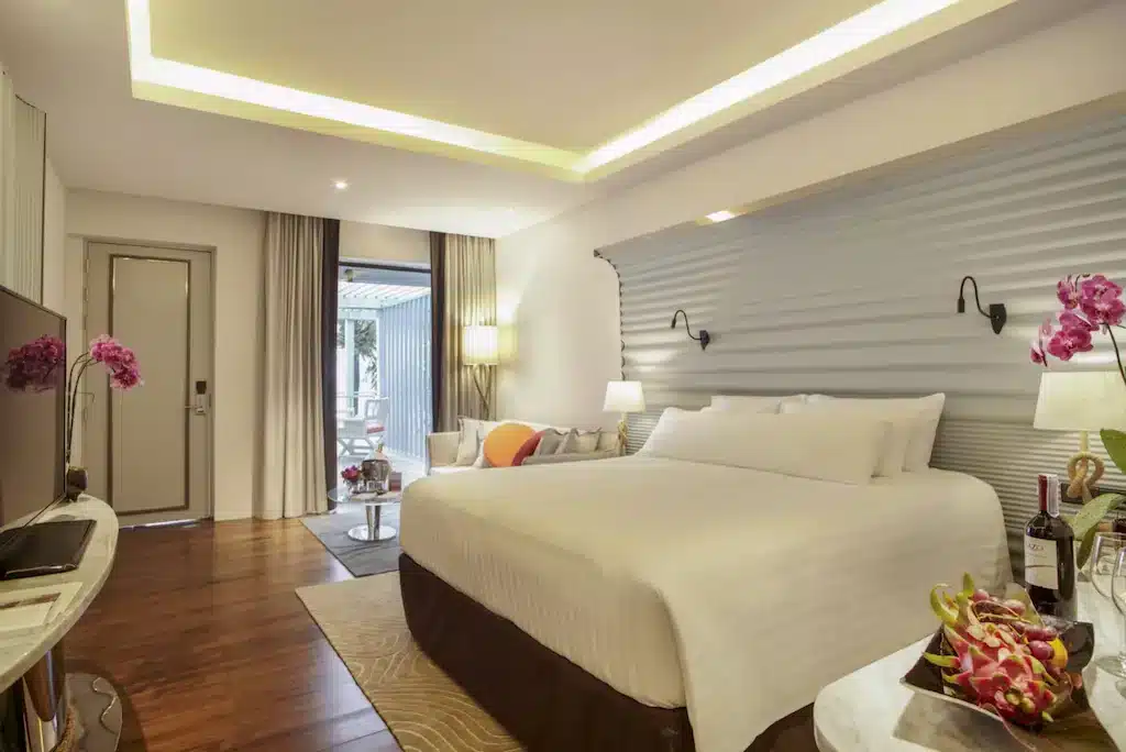 Modern hotel room with a large bed draped in white linens, a small sitting area with a coffee table and chairs, a wall-mounted TV, and a desk. The room features wooden flooring, contemporary light fixtures, and vibrant pink orchids on the table. Ideal for those exploring luxury beachfront resorts in Phuket.