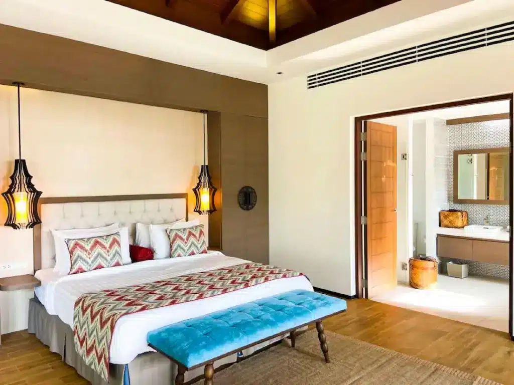 A spacious, well-lit bedroom features a large bed with a tufted headboard, chevron-patterned pillows, and a blanket. Two pendant lights hang on either side. A wooden bench with blue cushioning is placed at the foot of the bed. To the right, an open doorway leads to a bathroom with a sink and mirror, adding tranquility reminiscent of Big Buddha Phuket.