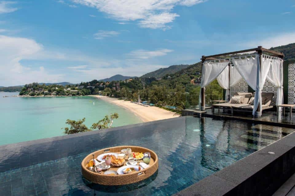 An infinity pool overlooking a scenic beach and ocean at one of the luxury beachfront resorts in Phuket. A floating bamboo tray with various breakfast items, including fruits and pastries, is in the pool. A white canopy bed with curtains is positioned beside it, and lush green hills are visible under a clear blue sky.