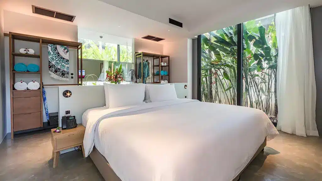 A modern bedroom features a large bed with white linens, flanked by wooden nightstands. Behind the bed are wooden shelves displaying decorative items and a hanging tapestry. Large windows with sheer curtains reveal lush green plants outside, reminiscent of the tranquil vibes at luxury beachfront resorts in Phuket.