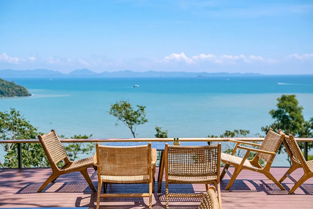 A scenic outdoor seating area with five wicker chairs arranged around a small table on a wooden deck. The seating overlooks a beautiful, expansive ocean view common to luxury beachfront resorts in Phuket. Trees frame the foreground, and distant mountains and a clear blue sky complete the idyllic scene.