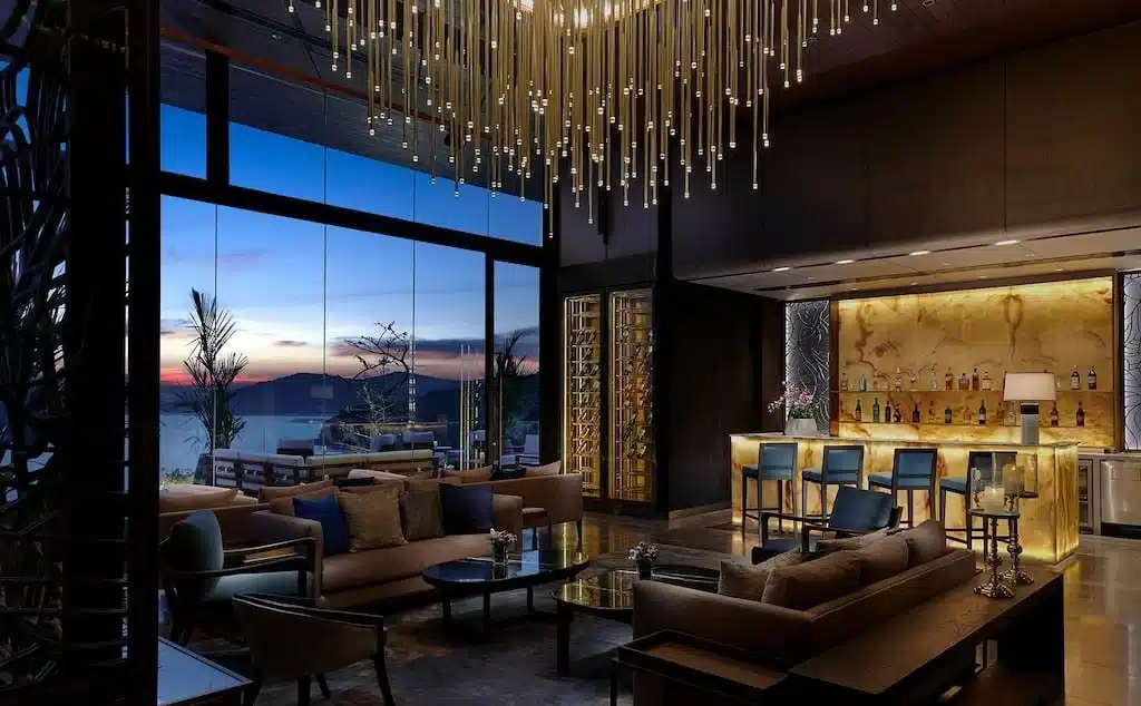 A modern, luxurious lounge found in one of the best hotels in Phuket near the beach features floor-to-ceiling windows revealing a sunset over the ocean. The room has stylish sofas, a sleek bar area with illuminated countertops, and a dazzling ceiling light fixture. The decor is elegant, with warm tones and contemporary design elements.