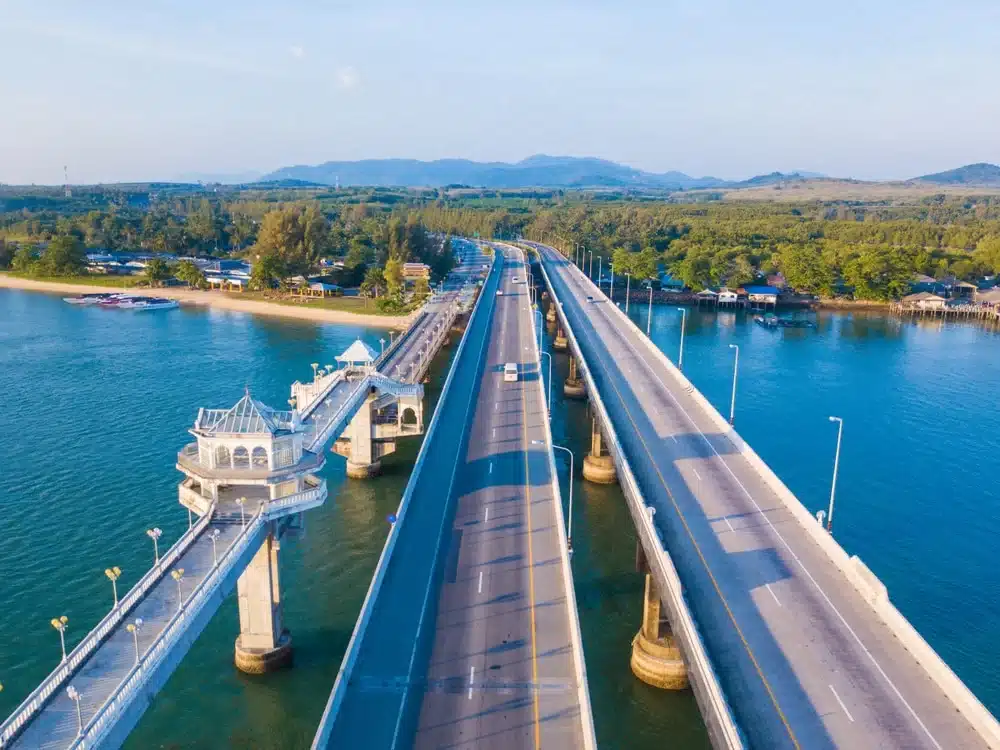 Aerial view of the Sarasin Bridge, a dual-carriageway bridge spanning over a body of water, linking two landmasses covered with trees and vegetation. An adjacent pedestrian bridge with a gazebo-like structure runs parallel to it. Mountains are visible in the distant background under a clear blue sky.