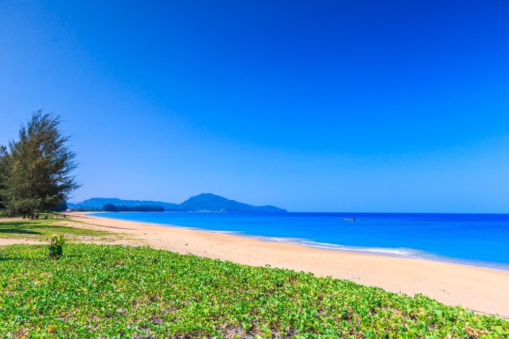 A pristine beach with golden sand stretches into the distance, bordered by verdant green vegetation. The turquoise ocean meets a clear blue sky on the horizon, with a single boat floating on the calm water. In the background, under the bright sunlight, a lush, tree-covered mountain rises beside Big Buddha Phuket.